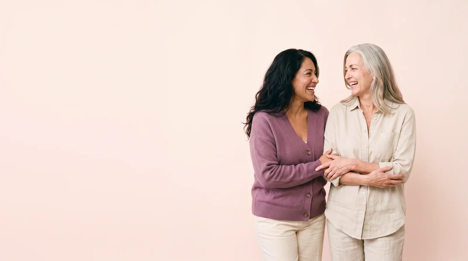 Two women arm in arm, laughing together against a pale blush backdrop.