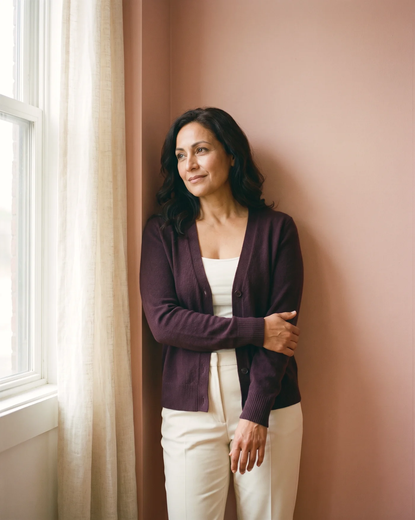 A woman in a plum cardigan leaning against a blush wall by a window, arms folded, smiling softly as she looks outside.